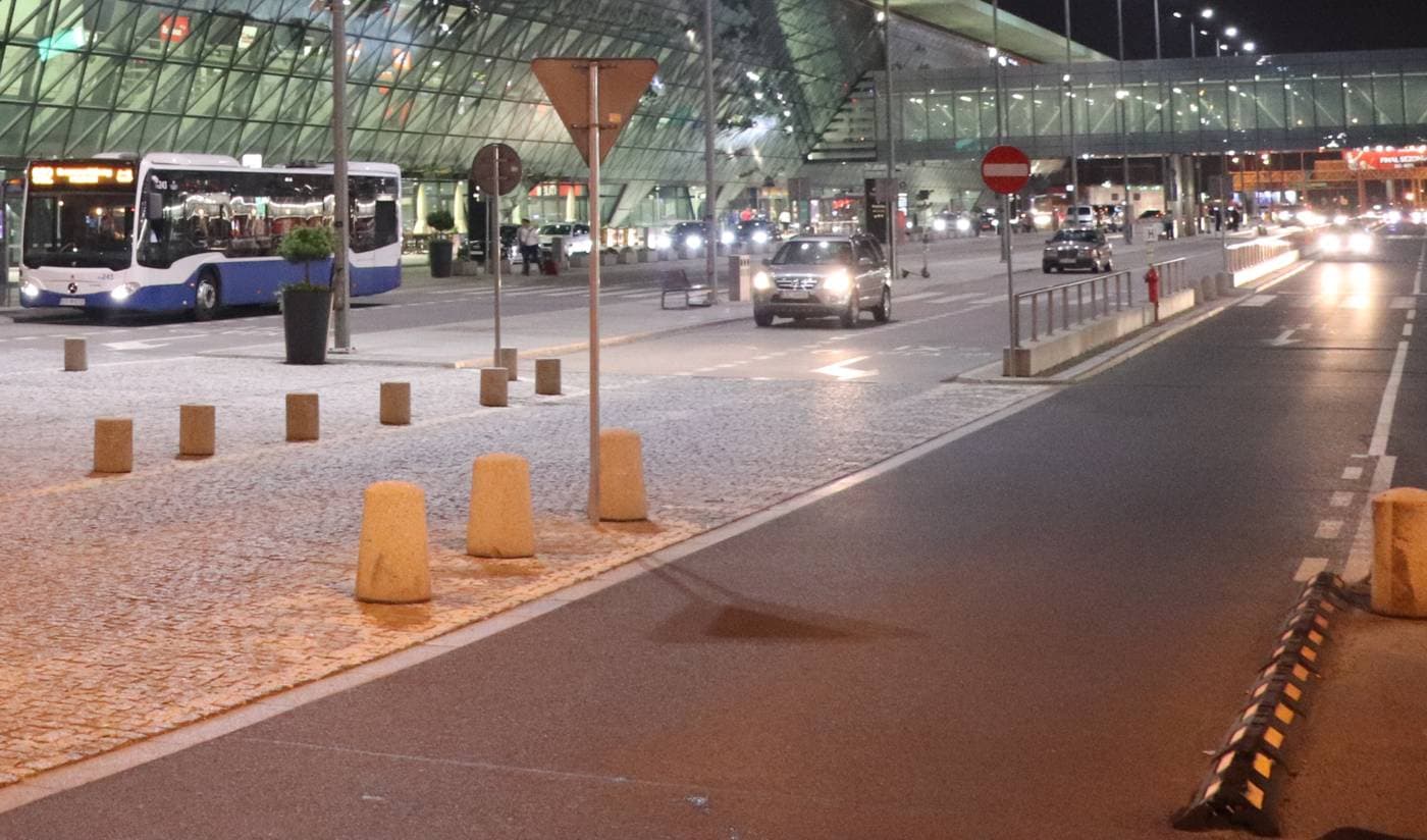 Podjazd pod terminal lotniska Kraków-Balice nocą: autobus przy krawężniku, bruk w świetle latarni i droga dojazdowa